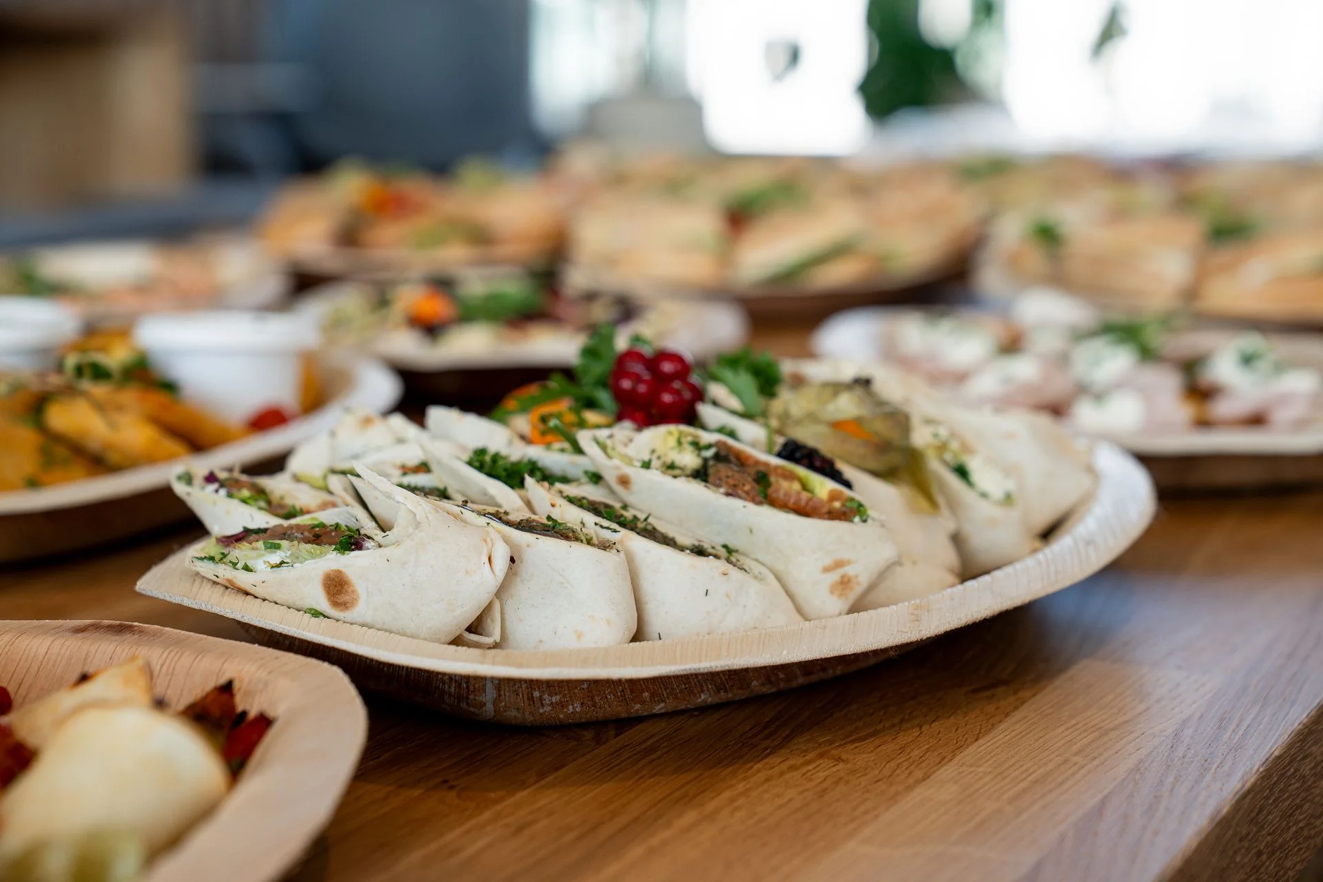 Platter of wraps with more sandwich trays behind on a wooden table